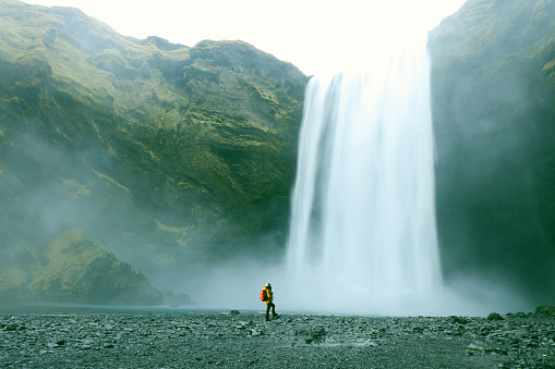 A female hiker is standing at a foot of a Skogafoss waterfall in Southern Iceland. Traveller looks very small in perspective with this majestic waterfall. Skogafoss is one of the best known and most popular waterfalls in Iceland and is here photographed on a cold windy day in winter of 2015. Longer exposure for the silky running water effect.
