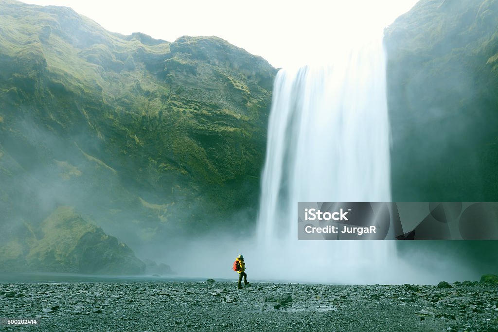 Hiker at Majestic Skogafoss Waterfall in Iceland A female hiker is standing at a foot of a Skogafoss waterfall in Southern Iceland. Traveller looks very small in perspective with this majestic waterfall. Skogafoss is one of the best known and most popular waterfalls in Iceland and is here photographed on a cold windy day in winter of 2015. Longer exposure for the silky running water effect. Large Stock Photo