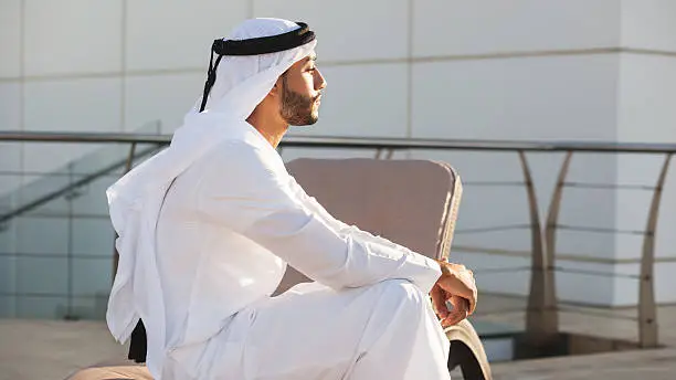 Muslim man is sitting on a beach chair on a rooftop of a hotel building and sunbathing his face in a golden sunlight. A peaceful moment in the break of the day. Background is a light grey building wall. Image contains copy space. Made in Dubai, UAE.
