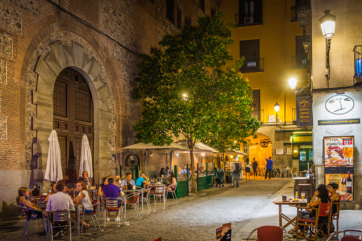 Madrid, Spain - July 11, 2015: People relaxing at the al fresco tables of a pavement restaurant bar illuminated by the warm lamp light of historic central Madrid, Spain's vibrant capital city.