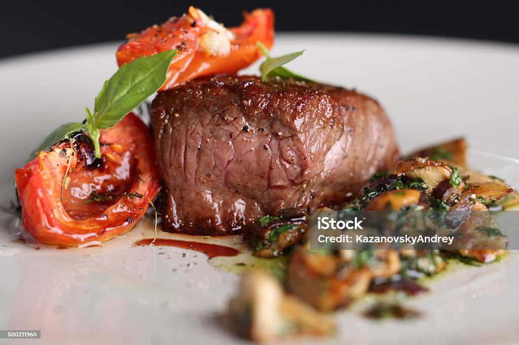 Roasted meat with tomatoes and mushrooms on a white plate Roasted meat with tomatoes and mushrooms on a white plate, dark background in the restaurant. Close up Gourmet Stock Photo