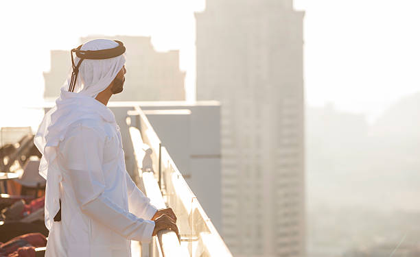 Traditionally dressed arab man is gazing into the horizons of a big city from a tall building's rooftop, resting his arms on the railings. The air is filled with haze, tall city buildings figure in the background. It's a hot evening filled with sunlight. Image contains plenty of copy space. Made in Dubai, UAE.