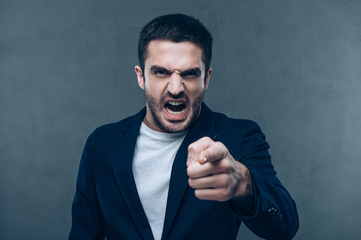 Furious young man looking at camera and pointing you while standing against grey background
