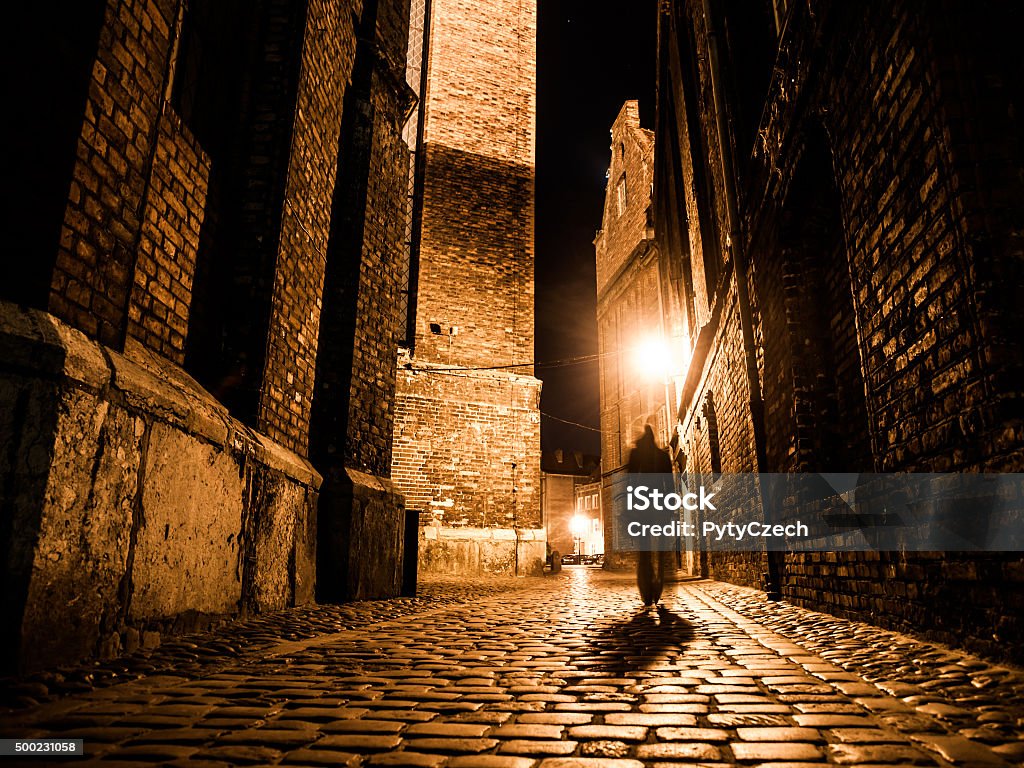 Illuminated cobbled street in old city by night Illuminated cobbled street with light reflections on cobblestones in old historical city by night. Dark blurred silhouette of person evokes Jack the Ripper. Jack The Ripper Stock Photo