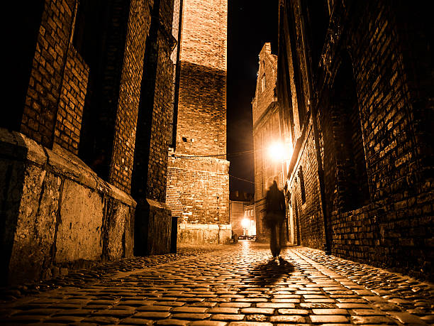 Illuminated cobbled street in old city by night Illuminated cobbled street with light reflections on cobblestones in old historical city by night. Dark blurred silhouette of person evokes Jack the Ripper. wall sidewalk city walking stock pictures, royalty-free photos & images