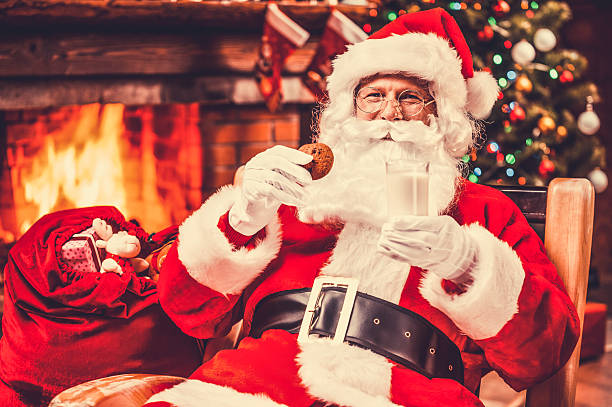 My favorite meal! Cheerful Santa Claus holding glass with milk and cookie while sitting at his chair with fireplace and Christmas Tree in the background calcium photos stock pictures, royalty-free photos & images
