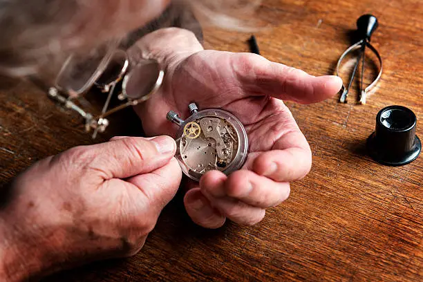 Close up  colour portrait of a watchmaker at work. In his hand you can see a watch that he trying to repair, he is wearing specialist magnifying glass and on this workbench are various watchmaking tools. The camera's  point of view is over his shoulder with the focus on the watch in his hand.