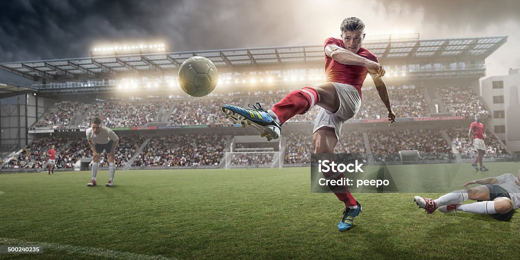 Soccer Player Kicking Ball A mid action image of soccer player kicking football, following a sliding tackle challenge from a rival player. The action takes place on a soccer pitch on a generic outdoor floodlit football stadium full of spectators under a stormy evening sky at sunset. All players are wearing unbranded generic kit.  Soccer Stock Photo