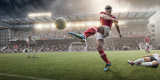 Soccer Player Kicking Ball A mid action image of soccer player kicking football, following a sliding tackle challenge from a rival player. The action takes place on a soccer pitch on a generic outdoor floodlit football stadium full of spectators under a stormy evening sky at sunset. All players are wearing unbranded generic kit.  soccer ball photos stock pictures, royalty-free photos & images
