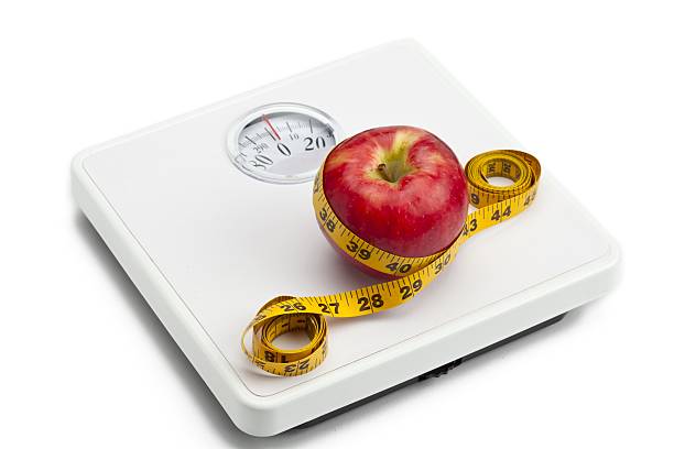 Apples and a measuring tape lie on the scales. Scales show zero. On brushed pine boards. Symbol of World No Diet Day.