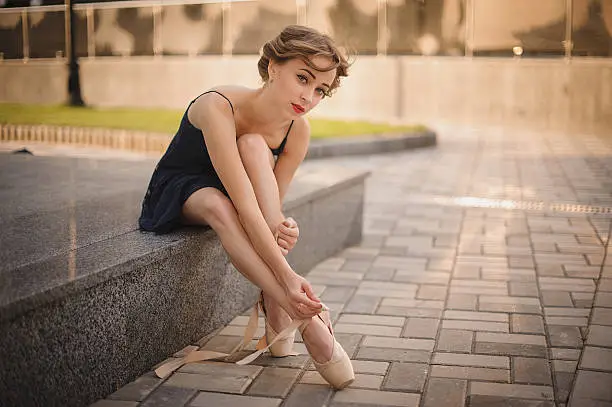 Slim young ballerina in a blackdress putting on pointe shoes. outdoor