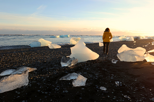 A woman is walking between big pieces of ice which are washed ashore near the Jokulsarlon glacial lagoon in Iceland. These icebergs are coming from a melting Vatnajokull Glacier and the whole coast is filled with them. This image is taken at sunset on a beautiful summer winter day.