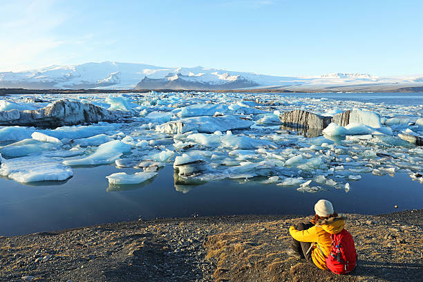 Female Hiker Looking at Icebergs at Jokulsarlon Glacial Lagoon Iceland Female hiker is looking at the drifting icebergs at the Jokulsarlon glacial lagoon in Southern Iceland on a beautiful sunny winter day in November of 2015. The traveller is wearing a bright yellow winter jacket and carrying a red backpack as she stands at the edge of a lake filled with ice formations from a melting Vatnajokull Glacier which can be seen in the background, jokulsarlon stock pictures, royalty-free photos & images