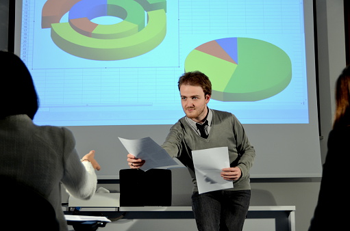 Teacher distributes text in a classroom, while pie charts are projected in the background.