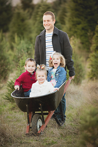 Father with 3 children in a wheelbarrow play for holiday photo in Christmas Tree Farm. Happy and smiling.