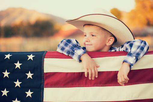 A young American boy dressed in cowboy western wear and cowboy hat displays the flag of the USA. He looks off camera and is smiling on the farm in Utah. 