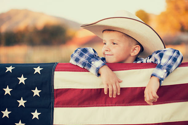 Young American Cowboy with US Flag A young American boy dressed in cowboy western wear and cowboy hat displays the flag of the USA. He looks off camera and is smiling on the farm in Utah.  texas cowboy stock pictures, royalty-free photos & images