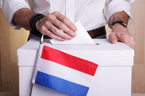 A man inserting a ballot to a ballot box. Dutch flag in front of it.