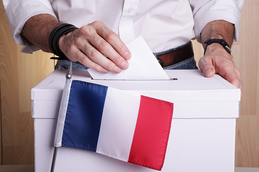 A man inserting a ballot to a ballot box.  French flag in front of it.