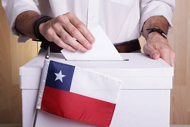 A man inserting a ballot to a ballot box.  Chilean flag in front of it.