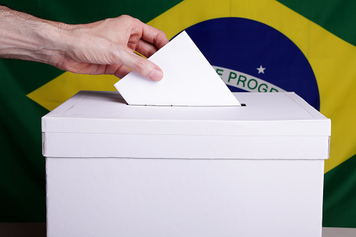 A man inserting a ballot to a ballot box.  Brazil flag in front of it.