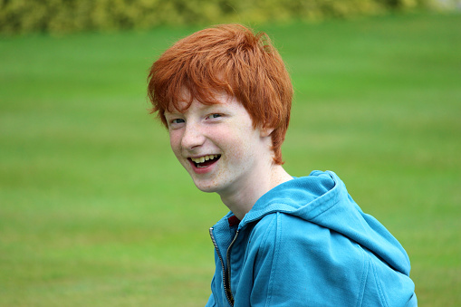 Photo showing a handsome young boy with short red hair, pictured smiling and laughing to himself as he sits in the garden.  The green lawn provides a natural, blurred background.