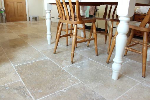 Photo showing a tiled floor in a real spacious kitchen diner, with large rectangular, filled travertine tiles laid lengthways in a brick pattern.  A pine table with painted white legs and beech chairs is pictured at the dining end of the kitchen.