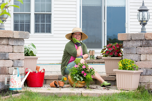 Grandmother potting up plants on her patio sitting on the brick step surrounded by fresh seedlings and flowerpots looking pensively at the camera with her chin on her hand with a friendly smile