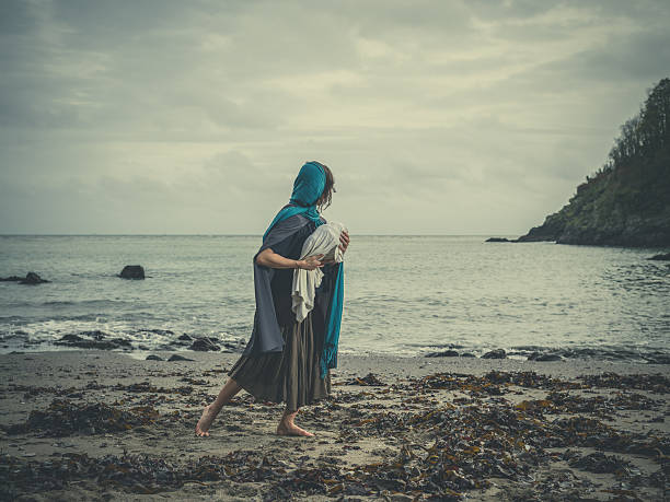 Vintage shot of woman on beach with baby Dramatic vintage filtered concept shot of a young woman on a beach clutching a baby wrapped up tightly in a blanket virgin mary photos stock pictures, royalty-free photos & images