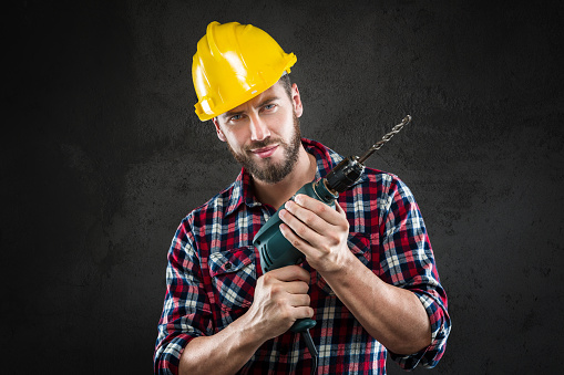 Portrait of attractive confident bearded workman with chequed shirt and yellow helmet, standing holding a drill, looking at camera, on a wall background