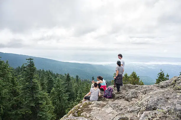 Canada:  A group of related backpackers look at a mobile phone mapping application and rest on the top of a mountain in a wilderness park at a popular viewpoint known as Dog Mountain.  The group is a real multi-ethnic and multi-generation extended family of mature adults and teenage children.  Wide angle view of the forest, mountain peak, and City of Vancouver in the background on a cloudy, overcast day.  Mount Seymour Provincial Park, North Vancouver, British Columbia, Canada.