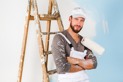 Portrait of handsome young smiling painter in white dungarees, splattered shirt and cap, leaning on a vintage ladder with paint roller