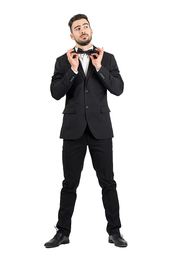 Young stubble groom adjusting bow tie looking up. Full body length portrait isolated over white studio background.