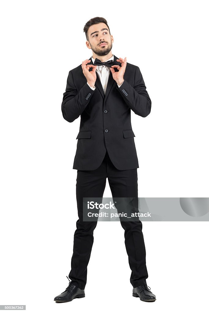Young stubble groom adjusting bow tie looking up. Young stubble groom adjusting bow tie looking up. Full body length portrait isolated over white studio background. Men Stock Photo
