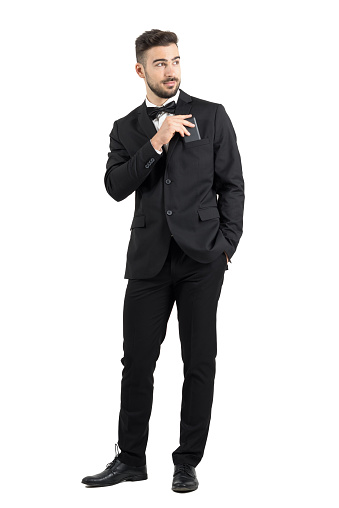 Relaxed cool handsome man in tuxedo with bow tie putting mobile phone in pocket looking away. Full body length portrait isolated over white studio background