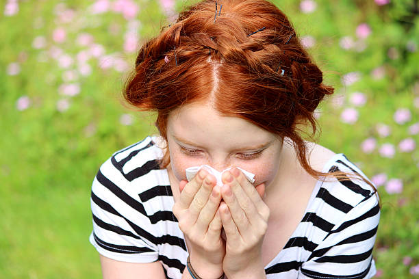 Girl in flower gardens with hayfever, blowing nose, sneezing, allergies Photo showing a young girl sat in a garden on a sunny afternoon, with pink hardy geraniums and grass seeds in the background.  She is pictured sneezing and blowing her nose, as she is suffering from hayfever and an allergy to pollen. blowing nose photos stock pictures, royalty-free photos & images