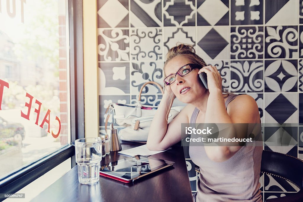 Urban woman working in a public cafe in the morning. Urban woman working in a public cafe in the morning. She is sitting on a stool, talking on mobile phone with digital tablet in front of her. Natural light coming from the window. She is wearing summer clothes. Horizontal shot with black and white pattern background. 40-44 Years Stock Photo