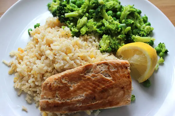 Photo showing a healthy meal consisting of a dry fried salmon fillet served with brown rice and red lentils, garden peas / frozen peas mixed with small pieces of broccoli and a slice of lemon. This dish is part of a healthy eating, low calorie diet plan.