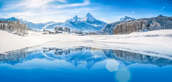 Panoramic view of beautiful white winter wonderland scenery in the Alps with snowy mountain summits reflecting in crystal clear mountain lake on a cold sunny day with blue sky and clouds.