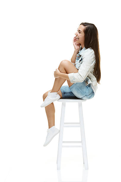 Side view of happy young woman sitting on stool Side view of happy young woman sitting on stoolhttp://www.twodozendesign.info/i/1.png jeans shorts women latin american and hispanic ethnicity stock pictures, royalty-free photos & images