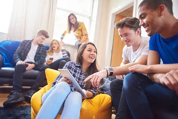 six young students sit around in their halls of residence , chatting and socialising with mobile phones and digital tablets. Three women and three men sit on sofas , beanbags and window ledges chatting and laughing .
