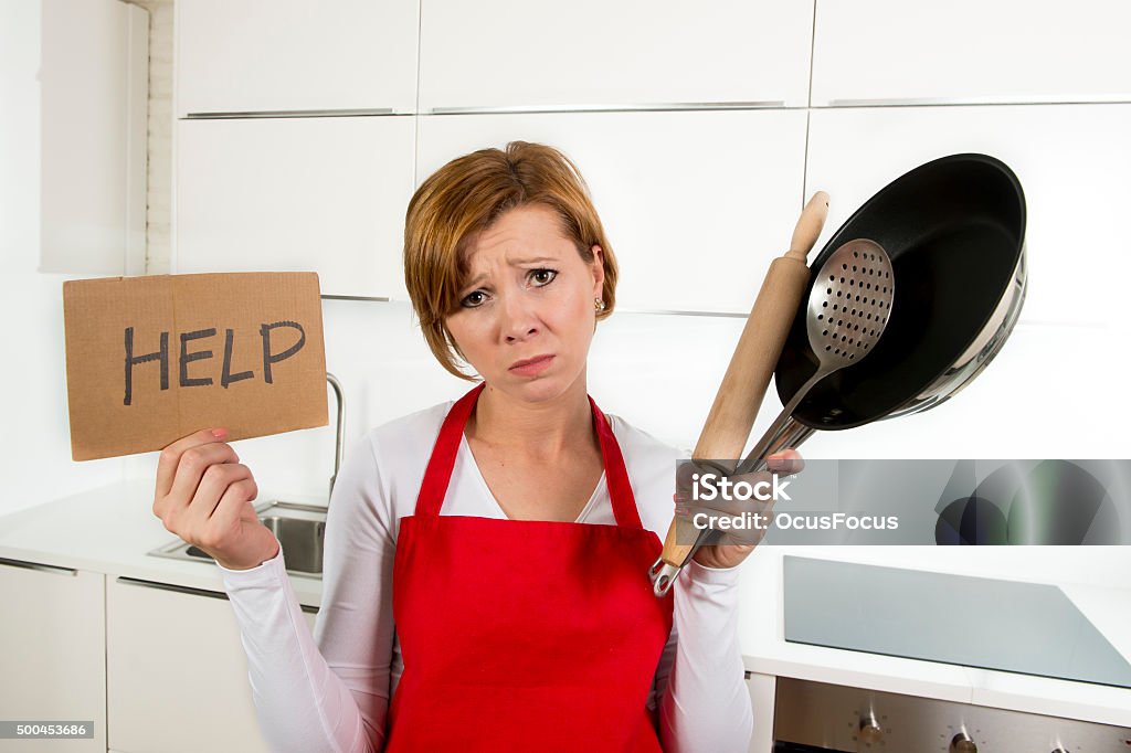 home cook woman holding cooking pan and rolling pin stressed young attractive home cook woman in red apron at domestic kitchen holding pan and household in stress with desperate and frustrated face expression in rookie amateur and inexperienced cooking Adult Stock Photo