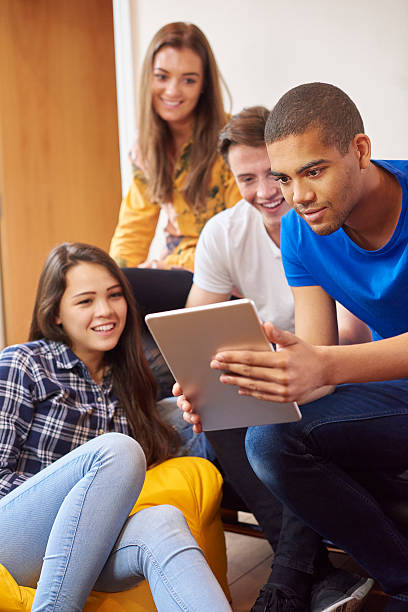 student tablet fun four young students sit around in their flat , chatting and socialising with  digital tablets. Two women and two men sit on sofas and beanbags chatting and laughing at what the young man in the foreground is doing on his digital tablet . multi ethnic group college student group of people global communications stock pictures, royalty-free photos & images