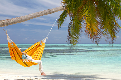 A young man wearing a straw hat is relaxing in a orange hammock that is hanged on a coconut palm tree. The white sand beach and the color of the water is turquoise and is an idyllic and unique location in The Maldives.
