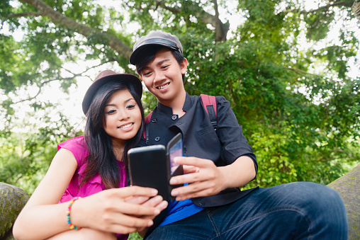 This is a horizontal, color, royalty free stock photograph of a young Balinese couple outdoors in Ubud, Bali. The Indonesian friends use a mobile phone to go online and look at social media. Photographed with a nIkon D800 DSLR camera.