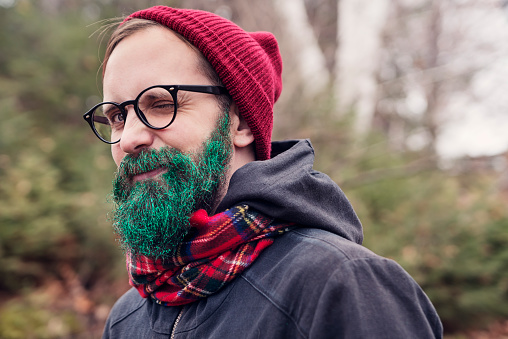 Can you be cooler than that? Hipster man ready for the holiday season with green glitter beard, a checked scarf and a red wool hat. He is winking at the camera. Kind of a hipster green Santa. Horizontal head and shoulder shot outdoors with copy space.