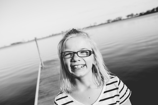 Black and white photo of a girl with glasses standing on a dock. 