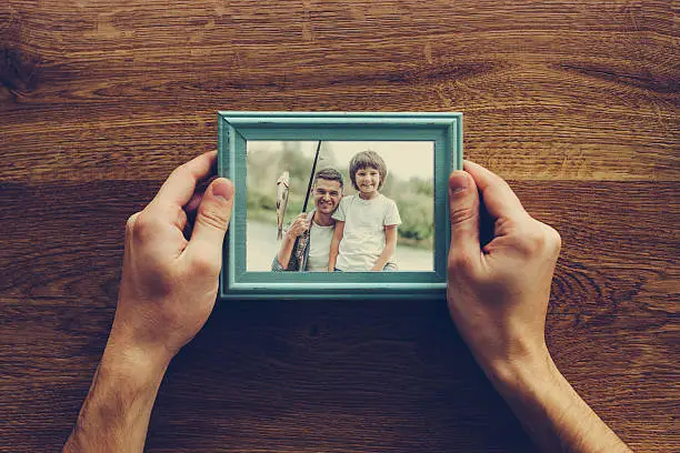 Close-up top view of man holding photograph of himself and his son fishing over wooden desk
