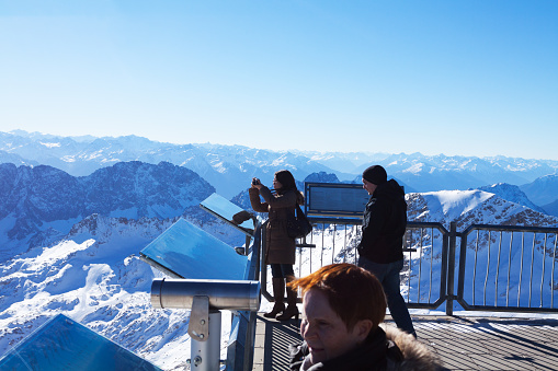 Grainau, Germany - November 21, 2012: Capture of some tourists on top of Zugspitze, German highest mountain in alps. A woman is taking photos. Other people are watching panorama.