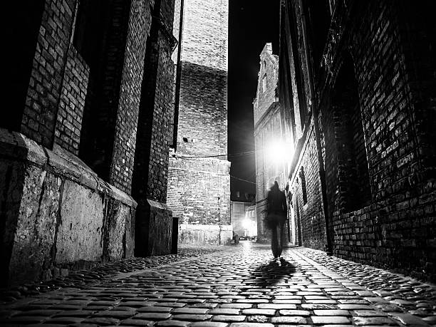Illuminated cobbled street in old city by night Illuminated cobbled street with light reflections on cobblestones in old historical city by night. Dark blurred silhouette of person evokes Jack the Ripper. Black and white image. cobblestone photos stock pictures, royalty-free photos & images